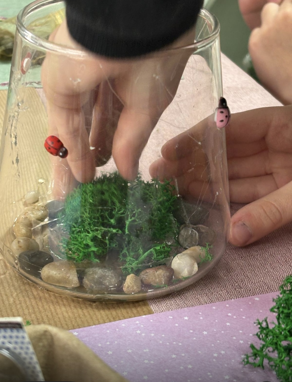 ידיים של ילד/ה מסדרות אבנים וצמחייה בתוך כוס זכוכית — יצירת טרריום בסדנת ArtyParty